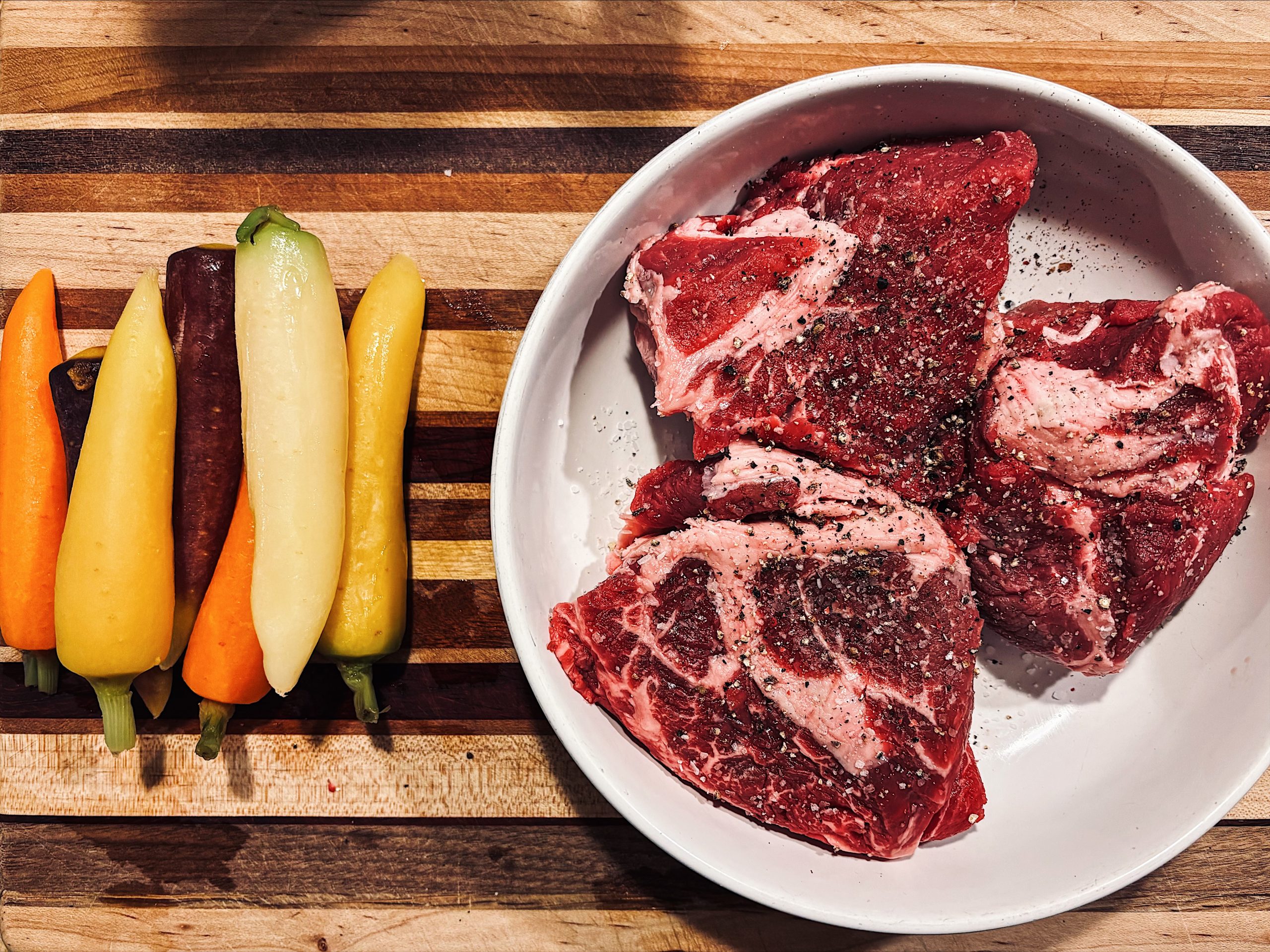 Raw steaks with pepper in a white bowl beside colorful whole carrots on a striped wooden board.
