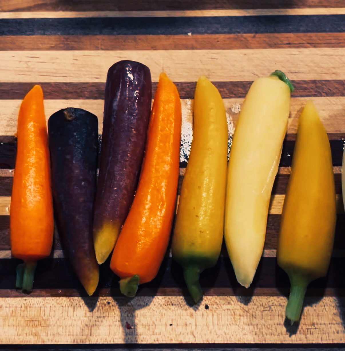 Six colorful carrots—orange, purple, yellow, pale yellow—lined up on a striped wooden cutting board.